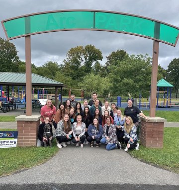 Occupational Therapy Students sit in a group under a park sign and smile.