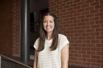 Kyla Reed, with long brown hair past her shoulders, wearing striped short sleeve shirt, stands in front of a brick wall and smiles.