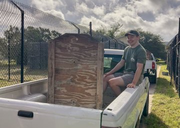 Marshall Hildreth in shorts, green t-shirt, and hat, sits in the back of a pick-up truck.