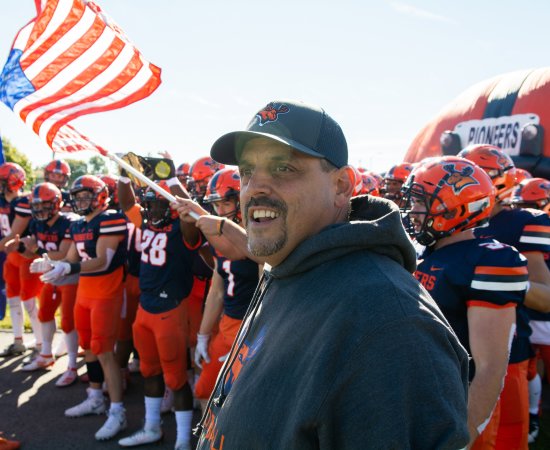 Coach Blaise Faggiano stands ahead of football players waving Pioneers flag