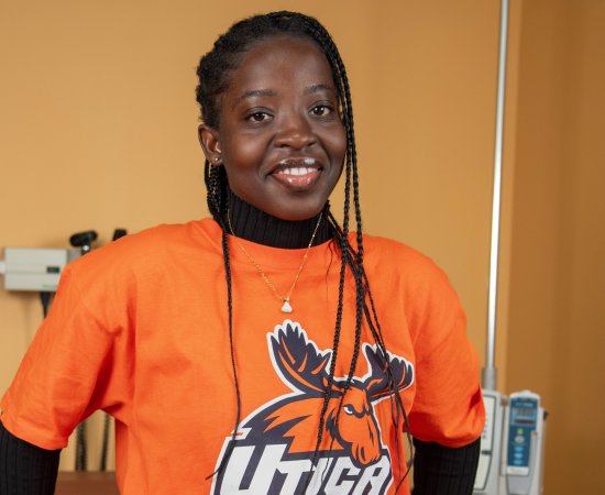 Lois Mensah, in an orange Utica t-shirt, stands against an examination table in the nursing lab.
