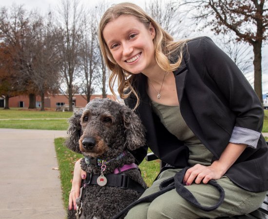 Young woman with blond hair wearing a black blazer and green shirt sits on a bench on campus with her arm around a black dog.