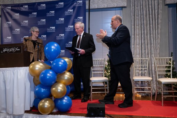 Dr. Thomas Rossi stands center on a platform stage, holding a plaque, as Provost Stephanie Nesbitt stands left and Genesis Group head Ray Durso to the right, both clapping.