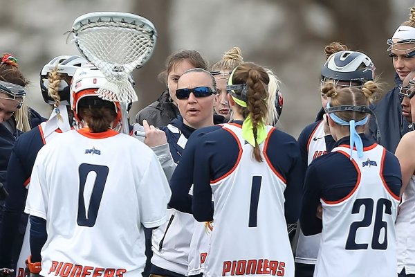 Kristin St Hilaire wears sunglasses and talks with a group lacrosse players on the field.