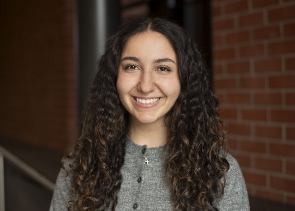Olivia D’Ambro, with long, dark, curly hair and a gray shirt, stands in front of a brick wall and smiles.