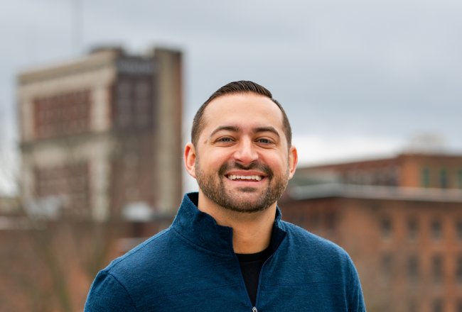 Thomas Armitage in a blue pullover, stands oustide with hands in pockets, smiling.