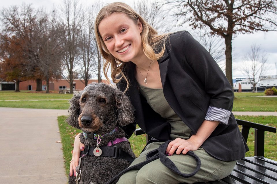 Young woman with blond hair wearing a black blazer and green shirt sits on a bench on campus with her arm around a black dog.